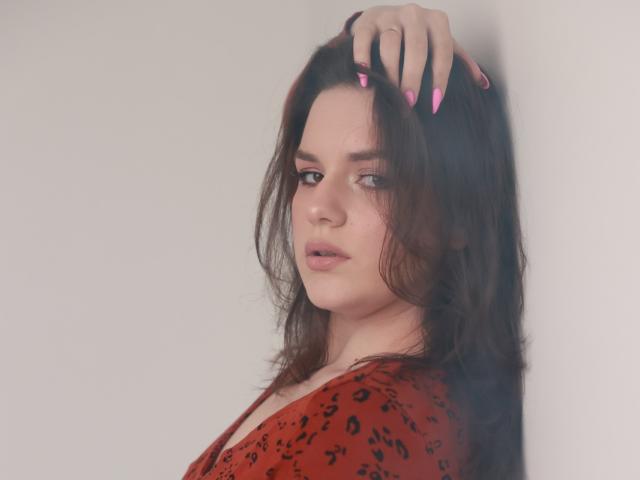 Dreia with pink nails, dark wavy hair, red leopard print top, leaning against white wall, looking over shoulder, soft pink lipstick, slightly parted lips, medium tits, subtle makeup.