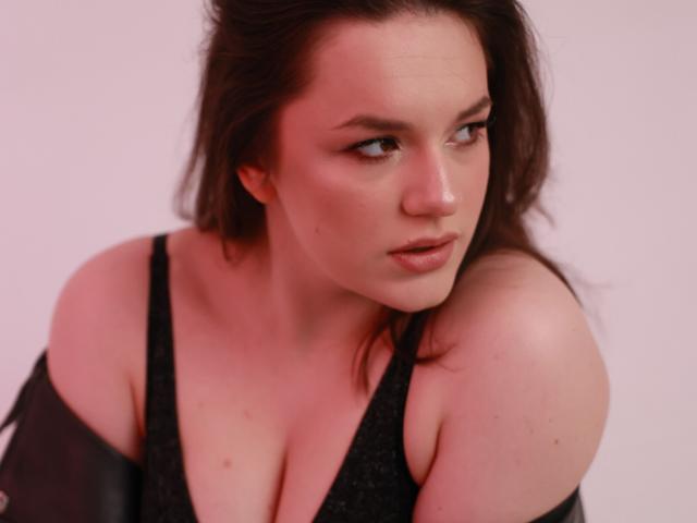 Dreia with fair skin and brown hair, looking to the side with subtle makeup, wearing a black sparkly top, showing her boobs and shoulders, with a black jacket off.