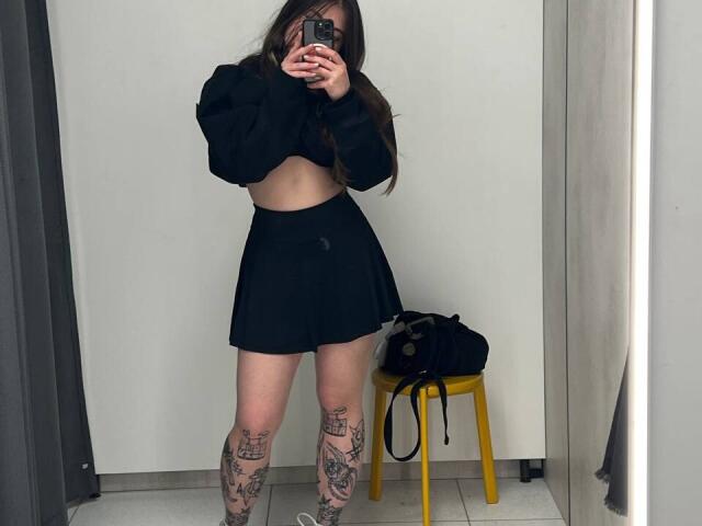 Mettis, avec de longs cheveux bruns, un crop top noir et une jupe noire, montrant ses cuisses tatouées et tenant un téléphone, se tient debout dans une pièce avec un tabouret jaune..