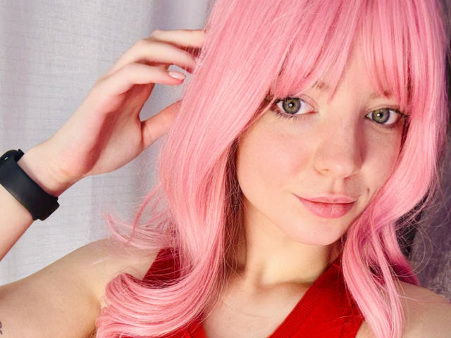 TeresitaBrugal with pink hair, green eyes, and red top, wearing a black wristband, has her hand near her head, showing off her freckled face and subtle smile.