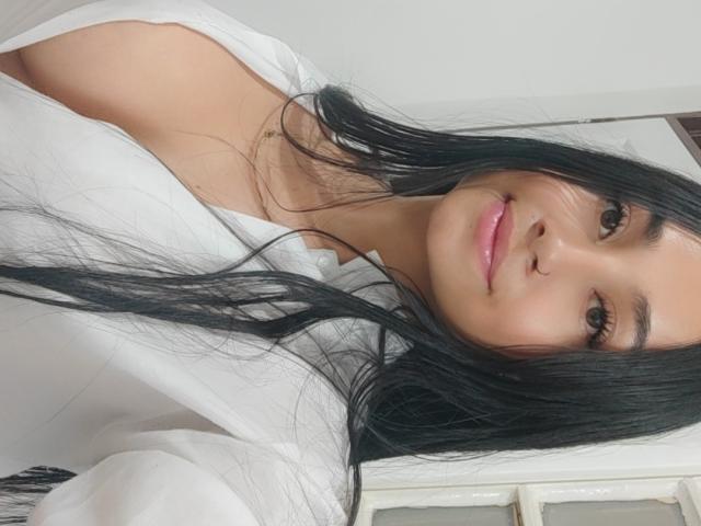 SexyCandyBabe69 with long black hair, pink lips, small nose ring, white shirt, and cute smile, showing bare shoulders and a hint of cleavage. Soft light, relaxed look.