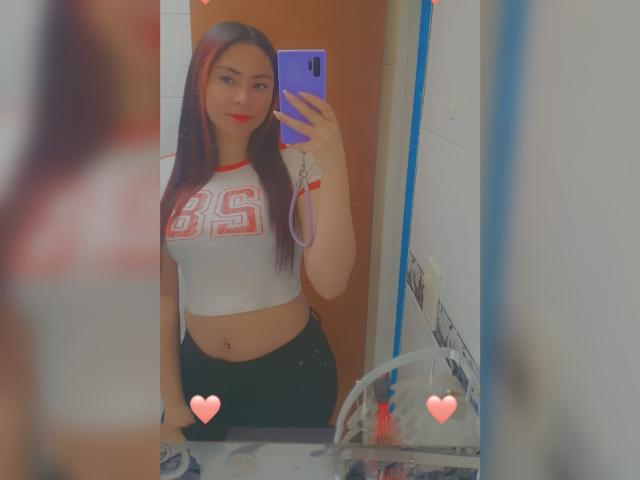 AnneDillinger with long red-brown hair, wearing a white crop top with red "BS" and black pants, taking a mirror selfie, showing her tits and a red lip.