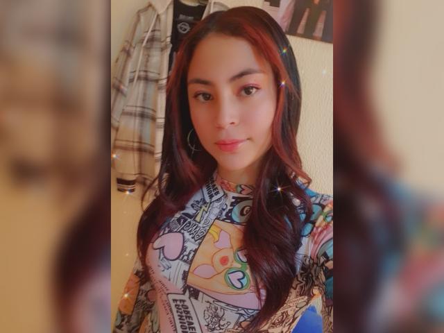 AnneDillinger with long red hair, wearing a colorful, patterned top, looking at the camera with a slight smile, small hoop earrings, soft lighting, and a gentle, cute face.