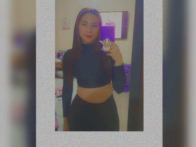 AnneDillinger, with long brown hair and red lipstick, wears a black long-sleeve crop top and black pants, holding a phone, showing her flat stomach and small ass.