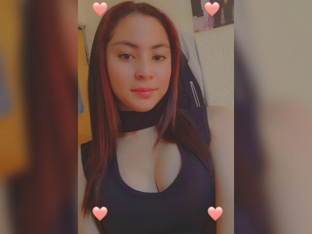 AnneDillinger has medium-sized tits in a black top, with straight dark red hair, a black choker, and a slight smile. Pink heart emojis are on the image.