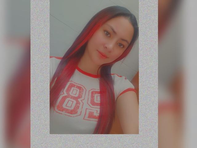AnneDillinger with long red and black hair, wearing a white shirt with red "88" on the chest, smiling softly, her cute face and small tits visible in the image.