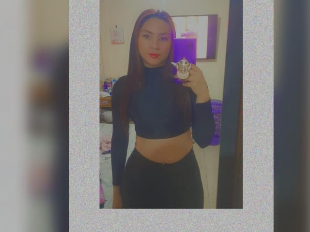 AnneDillinger with long brown hair, red lipstick, black crop top, and black pants, holding phone in mirror selfie, showing a slim, curvy, sexy figure with small, perky tits.