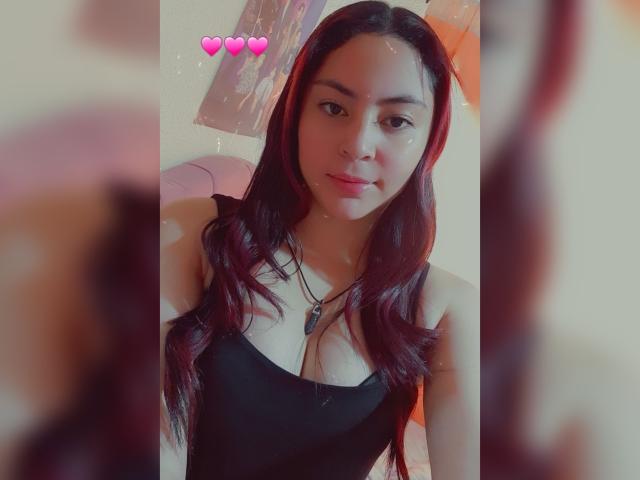AnneDillinger with long red hair, black tank top, necklace, and pink heart emoji, looking at camera with a slight smile, blurry foreground.