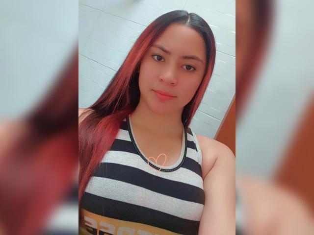 AnneDillinger with red and black straight hair, wearing a striped tank top, small heart necklace, and a subtle smile, showing her tits in a selfie.