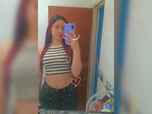 AnneDillinger with red and black hair, striped crop top, black pants, holding purple phone, mirror selfie, small heart sticker, bathroom, visible belly button piercing, slightly blurred.