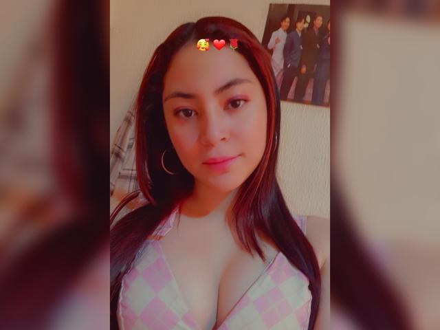 AnneDillinger with medium brown skin, long dark red hair, wearing a pink and white checkered top that shows her big boobs. She has a small heart emoji and red heart emoji above her forehead.