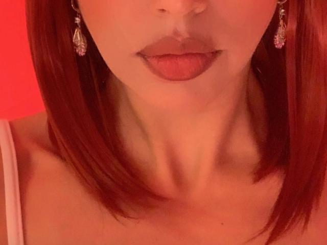 Os lábios vermelhos da BelladonnaLips estão ligeiramente entreabertos, revelando um vislumbre de sua língua. Ela tem cabelo ruivo, brincos rosa e um pescoço visível contra um fundo vermelho quente..