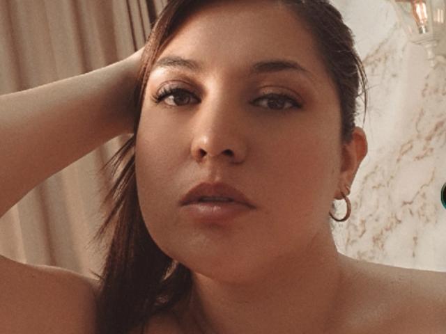PaulaAnd has brown eyes, slightly parted lips, and wears small hoop earrings. Her hand's in her brown hair, and she's looking directly at the camera.