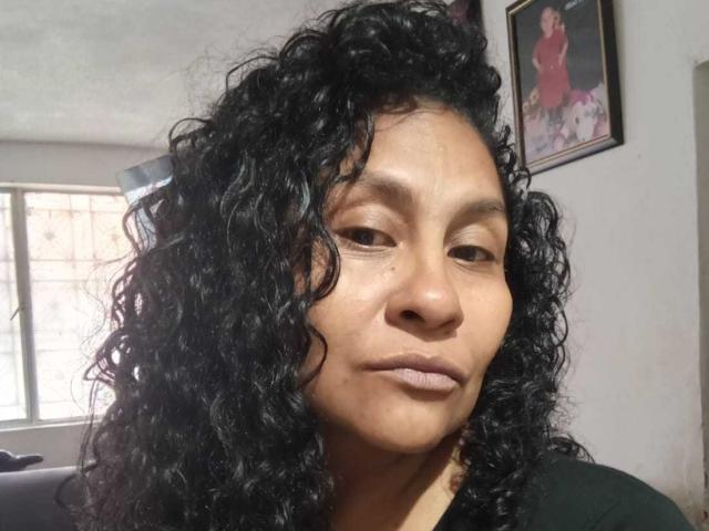 YessyForx ha i capelli neri e ricci, la pelle di un marrone medio e un'espressione neutra. Indossa una maglietta nera e dietro di lei, al muro, c'è una foto incorniciata..