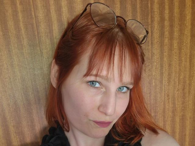 Ginger-Babe, una mujer atractiva con cabello rojo, ojos azules y gafas en la cabeza, tiene una ligera sonrisa, piel clara y lleva una blusa negra que deja al descubierto sus hombros..