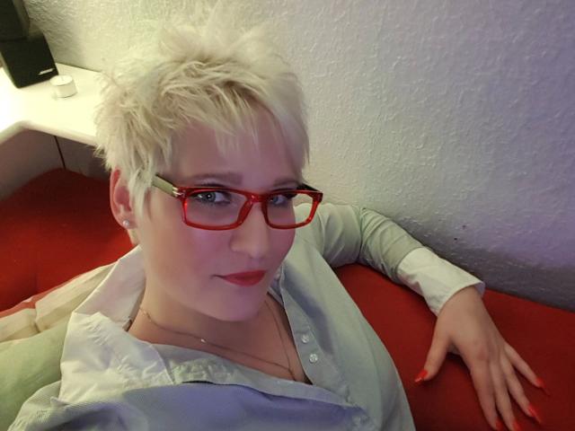 Ginger-Babe-atraente, com cabelo loiro curto e bagunçado, óculos vermelhos e unhas vermelhas, vestindo uma camisa verde clara, sentada em um sofá vermelho, olhando para a câmera..