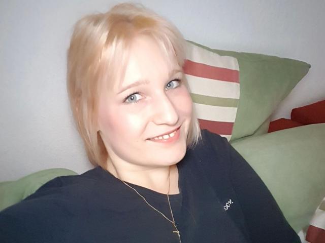 Una ragazza bionda, con occhi azzurri e un sorriso, indossa una maglietta nera e una collana d'oro, seduta su cuscini verdi a righe. Il suo seno è piccolo e i suoi glutei non sono visibili..