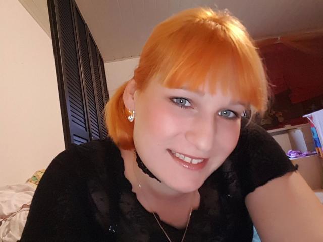 Ginger-Babe-sexy, com cabelo ruivo, olhos azuis e um colar preto, sorrindo para a câmera, vestindo uma blusa preta de renda, com brincos fofos..
