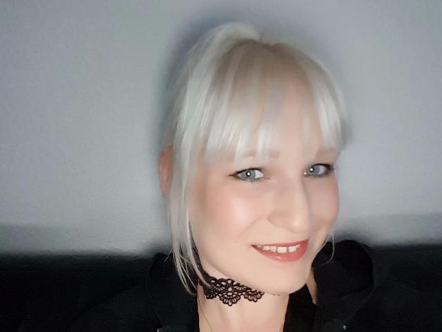 Una ragazza bionda platino, con occhi azzurri e un collare di pizzo, sorride alla fotocamera. Indossa una felpa nera, e il suo sedere non è visibile..