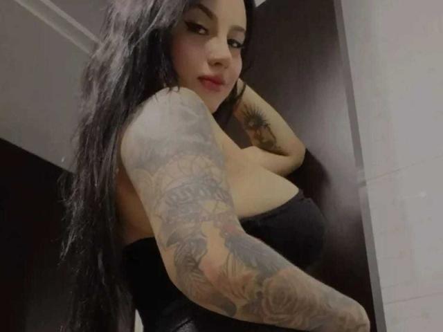 SamanthaLune with long black hair, tattooed arm, and black strapless top, looking over her shoulder, showing her tits, in a slightly blurred, indoor photo.