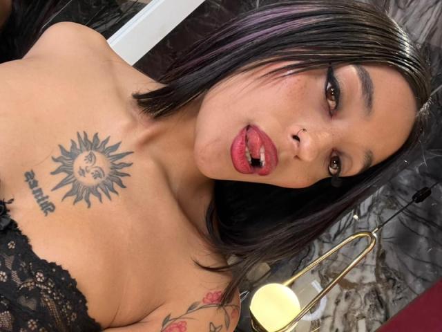 SalomeMille has black hair, purple highlights, and a sun tattoo with a face on her chest. She's topless with red lips, tongue out, and a nose ring.