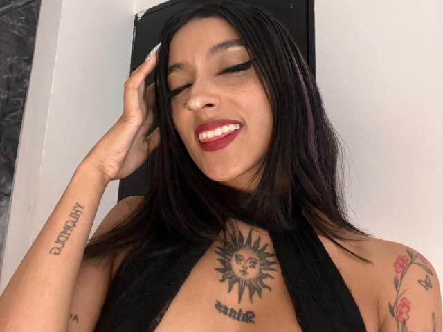 SalomeMille has dark hair, red lipstick, and a nose ring. She's smiling with eyes closed, showing tattoos on her chest and arm, wearing a black halter top.