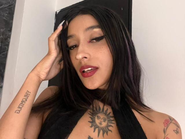 SalomeMille with dark hair, red lipstick, and black halter top, showing off sun tattoo on chest, "Bitch" tattoo on arm, and hand in hair. Her tits are visible.