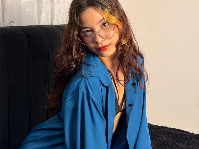 Vallerry69, com cabelo castanho encaracolado, uma mecha amarela e batom vermelho, usando óculos e uma camisa azul, sentada em uma cadeira preta, revelando um sutiã preto e o colo..