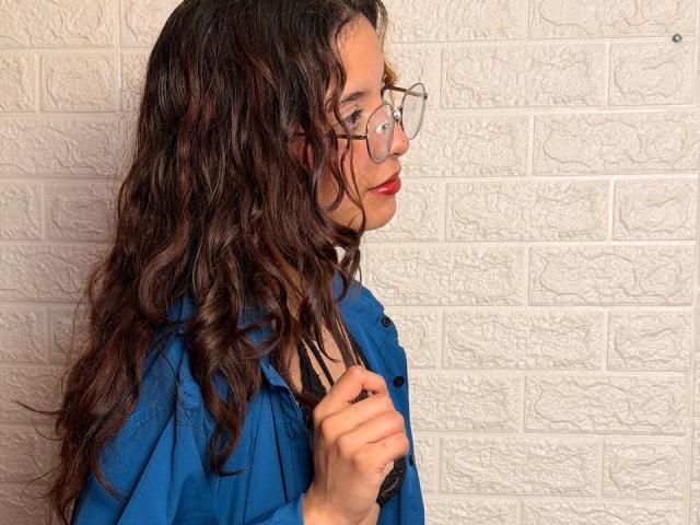 Vallerry69 with wavy brown hair, glasses, red lipstick, blue shirt, holding black bra, side profile, textured white wall, gentle smile, light skin, casual, confident.