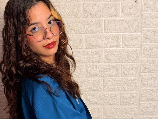 Vallerry69, avec des cheveux bruns bouclés, du rouge à lèvres rouge et des lunettes, portant une chemise bleue, regardant par-dessus son épaule, debout devant un mur de briques blanches texturées..