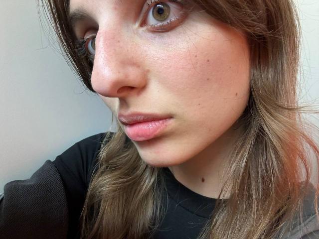 KarieMestler's face with light skin, brown eyes, pinkish lips, slightly freckled, brown wavy hair, wearing a black shirt, looking to the side with a subtle, calm expression.