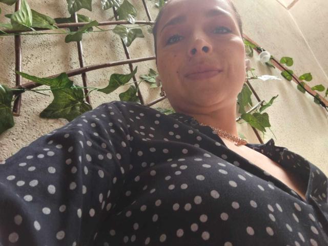 StangelarRivera, con una camicia nera a pois, una collana dorata e un sorriso discreto, guarda verso il basso la fotocamera. Sullo sfondo, rampicanti con foglie verdi. Ha la pelle marrone chiaro, capelli scuri, e un naso piccolo..