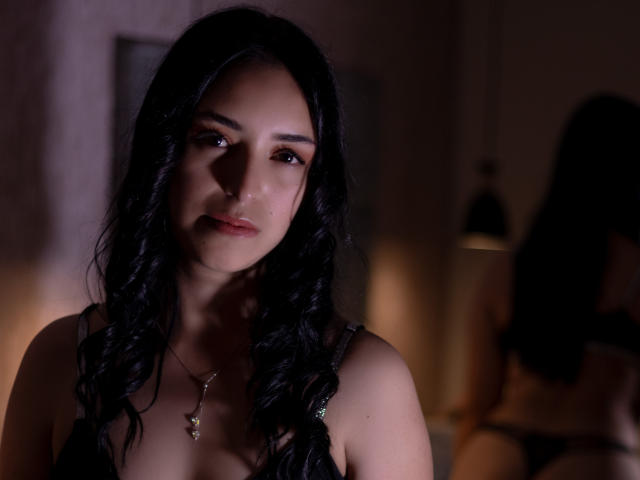 MartinaRey, com cabelo preto ondulado, olhos castanhos e um colar pequeno, vestindo um sutiã preto, em iluminação fraca, olhando ligeiramente para o lado, mostrando seus seios..
