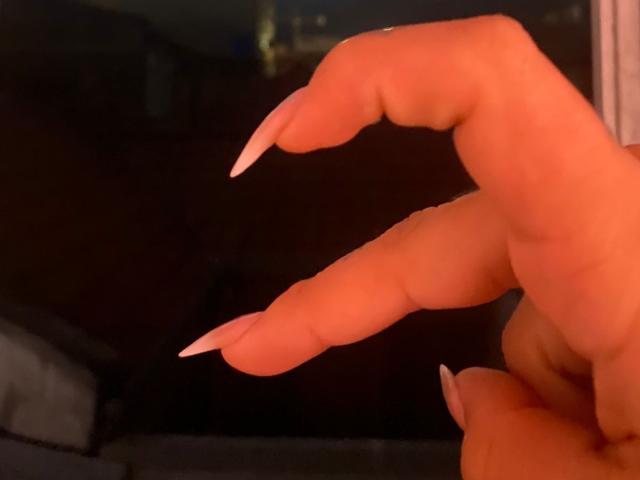 AmaraKazumi's hand with long, pointed, nude-colored fingernails, showing three fingers in the foreground. The skin is smooth and slightly glossy.