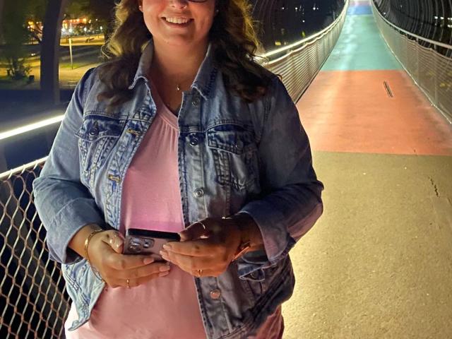 DirtyQueen-hot with curly brown hair, wearing a blue denim jacket and pink shirt, holding a smartphone, standing on a bridge at night, smiling. Her hands are on the phone.