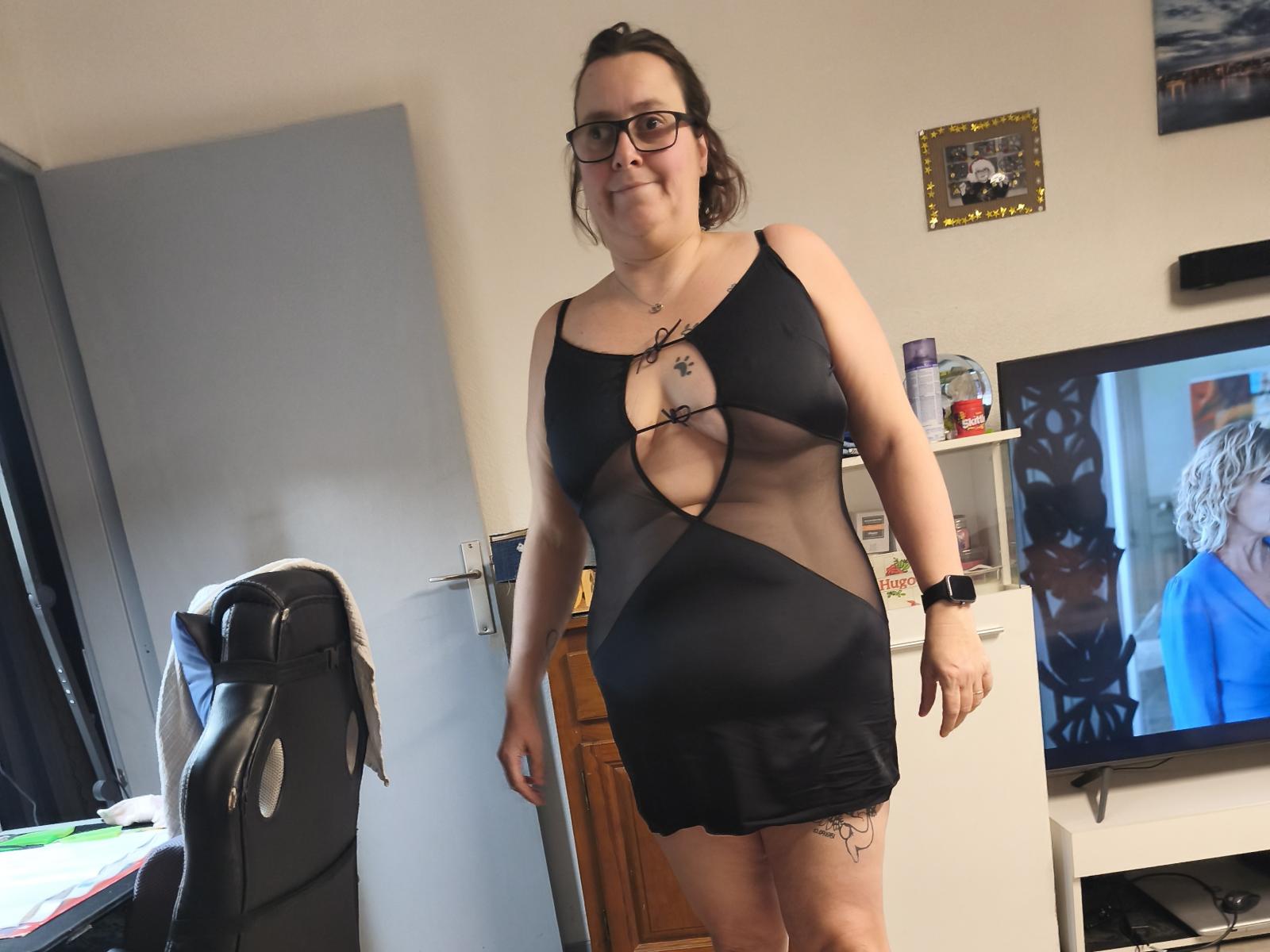 AngesEtDemons69, uma mulher corpulenta com óculos, veste um vestido preto transparente com um recorte em formato de chave, que revela seus seios e uma tatuagem na coxa. Ela está em pé em um quarto..
