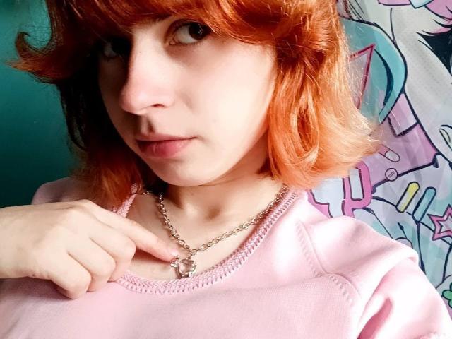 Mireille Spotwood mit rot-orangefarbenem Haar, heller Haut und einem pinken Hemd, die eine Halskette mit einem violetten Stein berührt und leicht zur Seite schaut. Ihre Brüste sind klein, und sie ist süß..