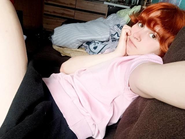 Mireille Spotwood mit roten Haaren, einem pinken Top ohne Ärmel, schwarzen Shorts, liegt auf einem braunen Sofa, die Hand in der Nähe des Gesichts, blickt in die Kamera, blasse Haut, entspannt, ein Arm ausgestreckt, ein Bein angehoben..
