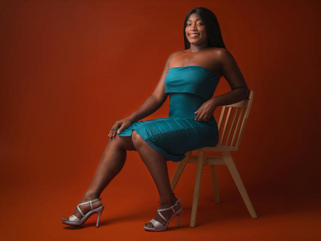 NayaKarter sits on a wooden chair, wearing a teal strapless dress, showing her ass, with dark skin, long black hair, and silver high heels, smiling.