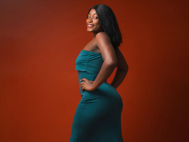 NayaKarter, dark-skinned with straight black hair, smiling, wearing a tight teal strapless dress, standing sideways, hands on hips, showing her ass and small, perky tits.