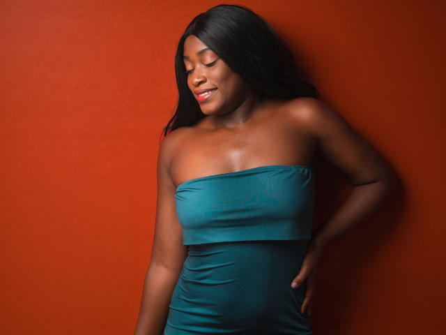 NayaKarter with dark skin, black hair, and closed eyes, wearing a teal strapless dress, hands on hips, smiling, red background, looking down, showing her boobs and ass.