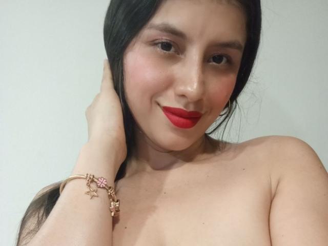MadissonFontaine with red lipstick, black hair in a braid, hand on neck, wearing a pink bracelet, topless, showing her tits, smiling, fair skin, against a white background.