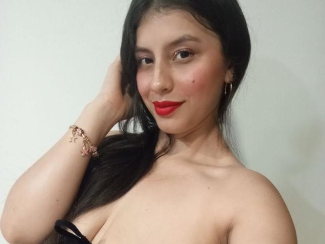 MadissonFontaine has black hair, red lipstick, and is topless with visible tits, wearing a bracelet, against a white background, smiling with a hand in her hair.