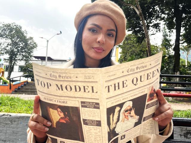 Jessica Fiore, com cabelo escuro, lábios rosados e um chapéu bege claro, segurando uma revista "Top Model The Queen", que mostra fotos de modelos, incluindo uma com um lenço na cabeça, em um ambiente externo..