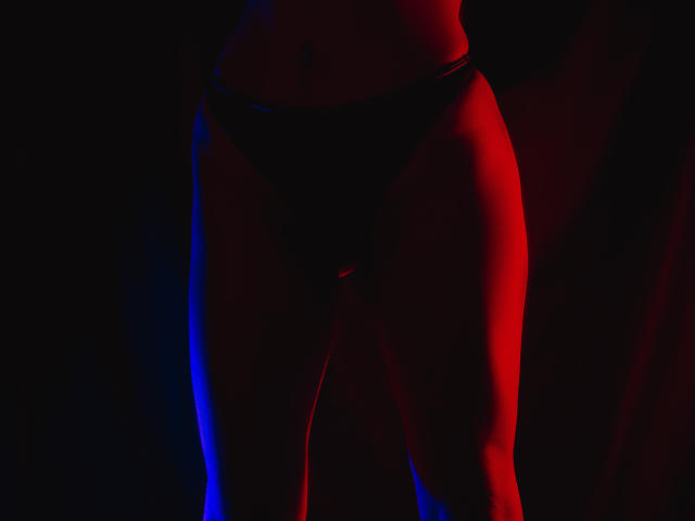 Helens schlanke, trainierte Beine mit schwarzen Unterhosen, hervorgehoben durch rotes und blaues Licht. Ihre Vulva ist durch den Stoff hindurch subtil sichtbar. Dunkler, schattiger Hintergrund..