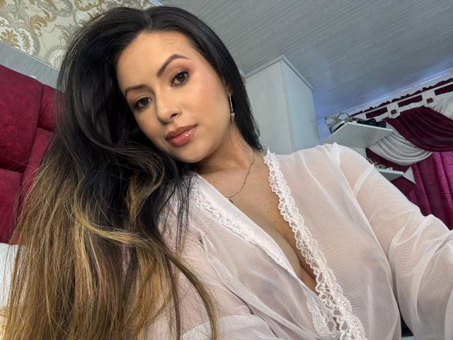 HannaRobertss, avec de longs cheveux noirs et blonds dégradés, portant une robe blanche en dentelle transparente, laissant entrevoir ses seins à travers le tissu, dans une chambre avec des rideaux rouges et blancs..