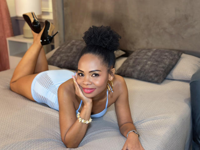 Chantal Haneke è sdraiata su un letto, indossa un abito leggero color azzurro con rete, tacchi neri e orecchini dorati, con i glutei sollevati e le mani sul viso, sorridendo..
