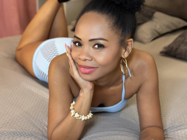 ChantalHaneke with dark skin, black hair in a bun, smiling, lying on a bed, wearing a blue mesh top, gold earrings, and a shell bracelet, resting on her cheek.