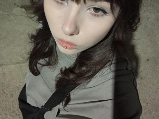 Eilene Szala tiene la piel pálida, el cabello oscuro y ondulado, y tiene el labio inferior perforado. Lleva una blusa gris y negra, y mira hacia arriba con los ojos entrecerrados..