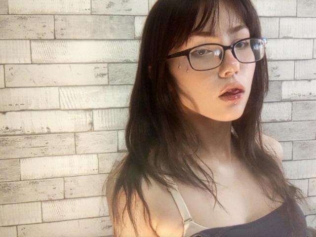 Eilene Szala, mit Brille, dunkelbraunem Haar und einem leichten Schmollmund, trägt ein schwarz-weißes Oberteil und zeigt dabei kleine Brüste vor einer weißen Backsteinmauer..